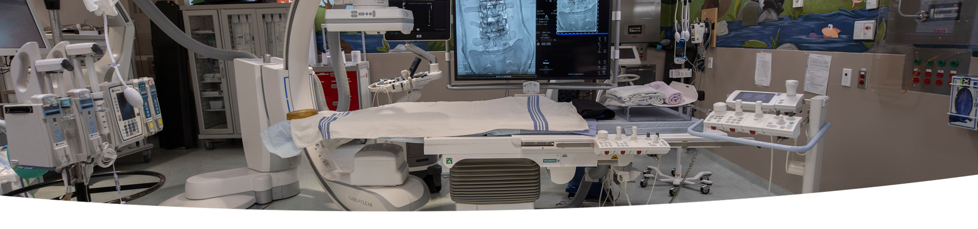 A room with a Single Plane Digital Angiography unit. There is a  variety of hospital equipment in the background.