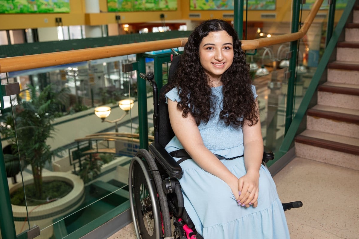 Melika sitting in a wheelchair and smiling for a photo on a staircase overlooking the Atrium Fountain.