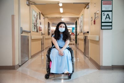 Melika sitting in a wheelchair in the middle of a hospital hallway. A sign on the wall says "5A".