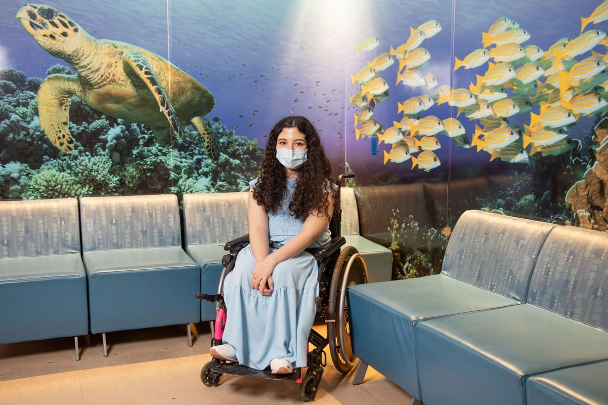 Melika sitting in a wheelchair in a waiting room area. The walls are decorated with wallpaper of animals in the ocean, such as sea turtles and fish.