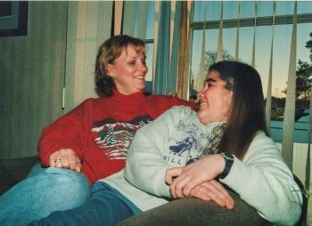 Kristina and Linda smile at each other, sitting closely on a couch in front of a window.