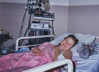 Kristina smiles at the camera while lying in a hospital bed.