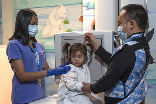 A child wearing a protective gown for X-rays while two staff in face masks prepare her for an X-ray test.