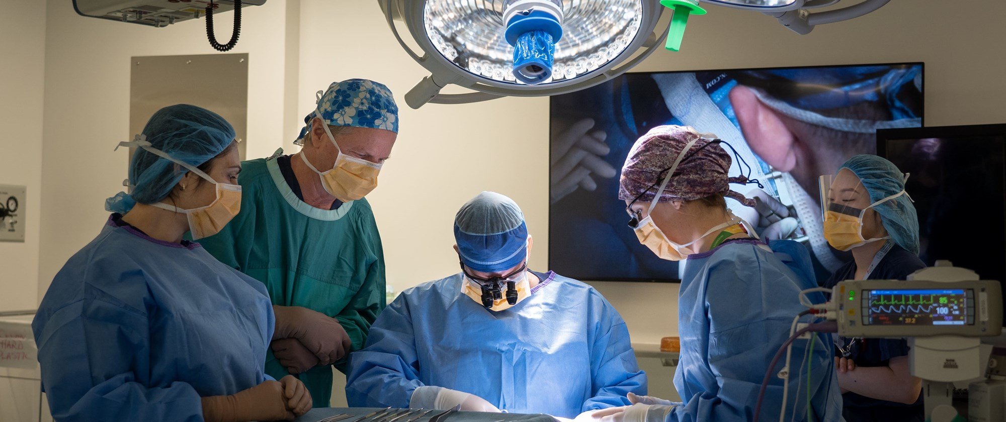 4 surgeons and assistants at the operating table with a digital display in the background and a bright light overhead