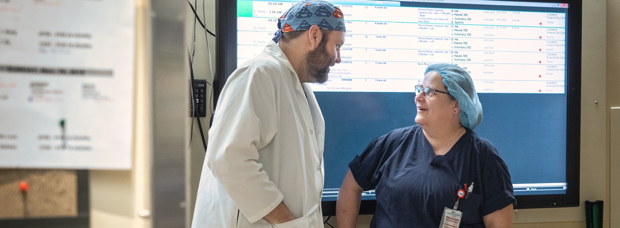 Two SK staff in PPE talking in a hallway in front of a digital chart display