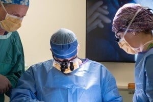 4 surgeons and assistants at the operating table with a digital display in the background and a bright light overhead