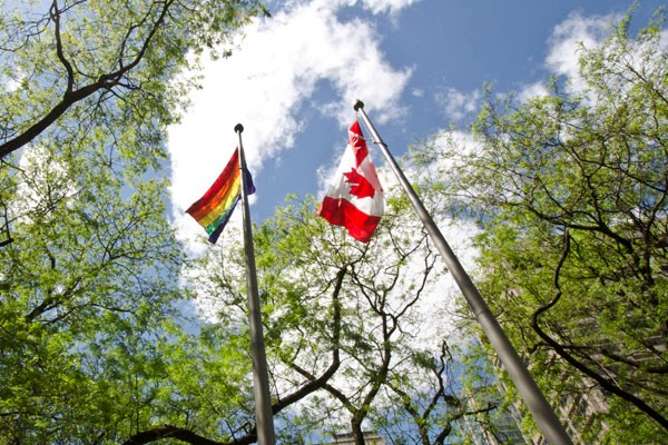 Rainbow pride flag and Canadian flag each flying from a flag pole. Photo taken from below.