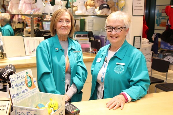 Two women wearing matching blue aprons stand behind a shop counter.