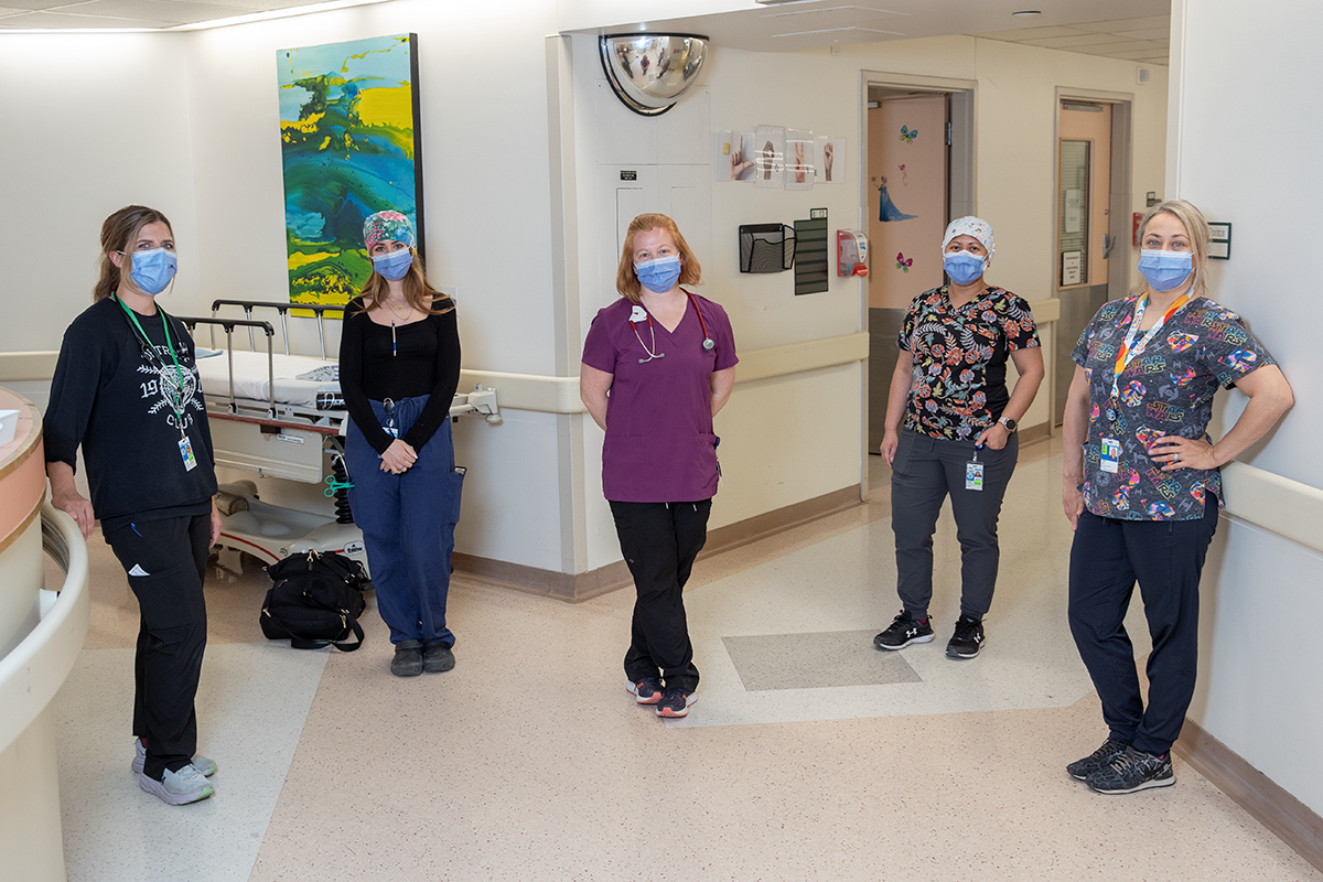 Five women stand distanced, wearing masks in a hospital hallway.