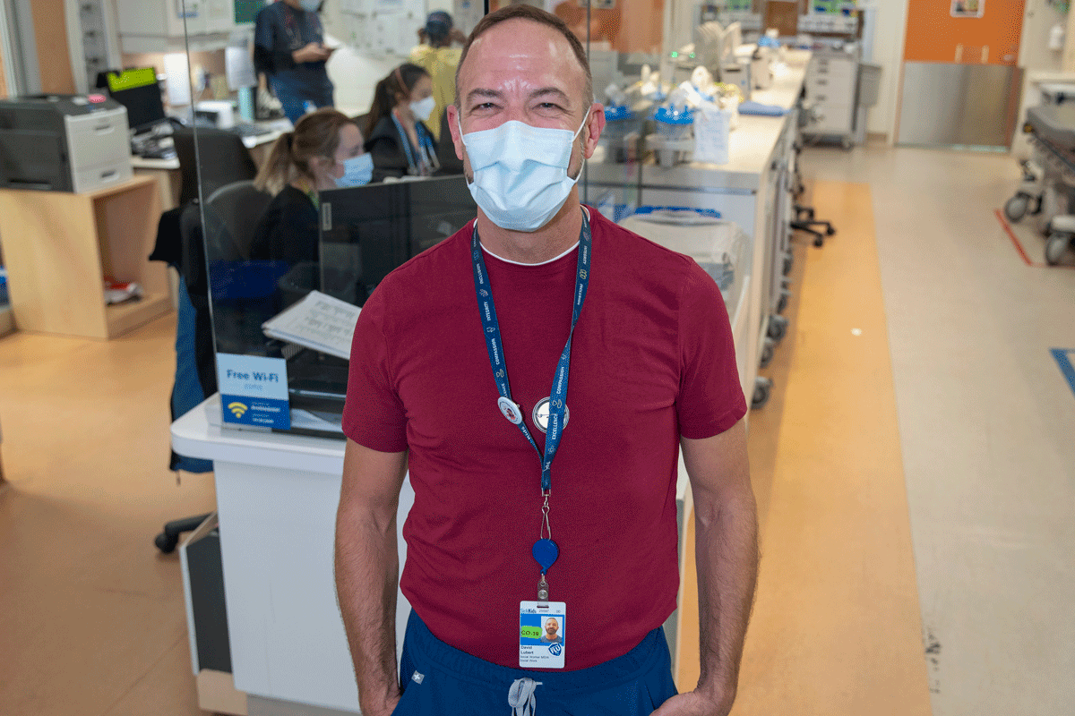 Social worker posing for the camera in front of the nursing station, hands in pockets, with Emergency Department in the background.