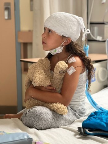 A girl sitting on a hospital bed while holding a teddy bear on her lap.