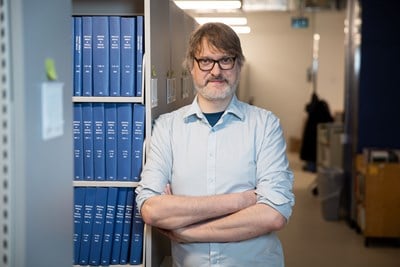 Bearded man in light blue shirt standing with crossed arms beside shelves of blue "American Journal" bound volumes in library.