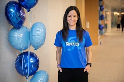 Krista Cardamone wearing a blue SickKids t-shirt standing in a hallway beside blue balloons.