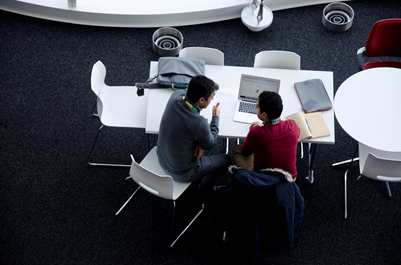 Two staff members having a discussion while seated at a table with a laptop and notebook open