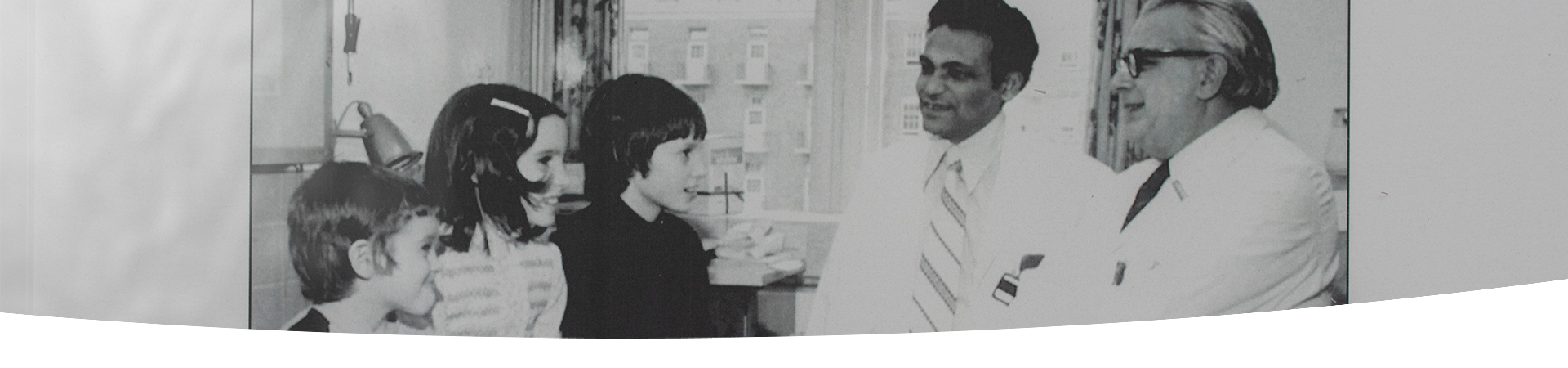 Black and white photo of Dr. Bibudhendra (Amu) Sarkar and Dr. Andrew Sass-Kortsak speaking with three children with Wilson disease