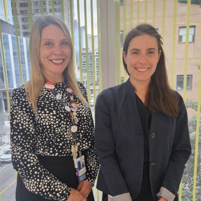 Two people standing indoors in front of large windows with a city view, smiling at the camera.
