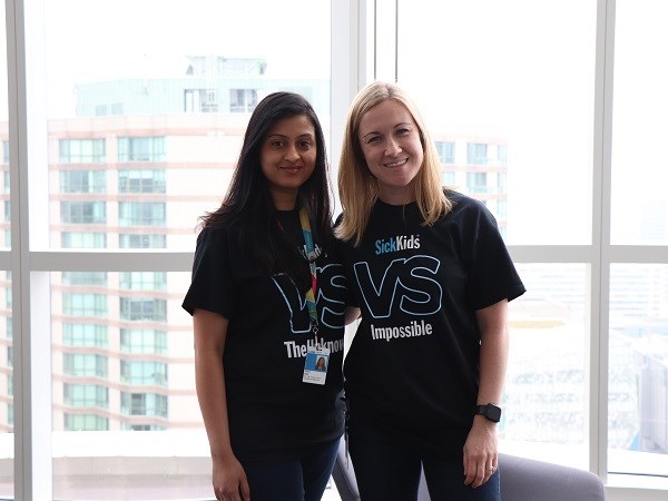 Two women stand side by side in front of a window with skyscrapers in the background. They are wearing SickKids t-shirts.