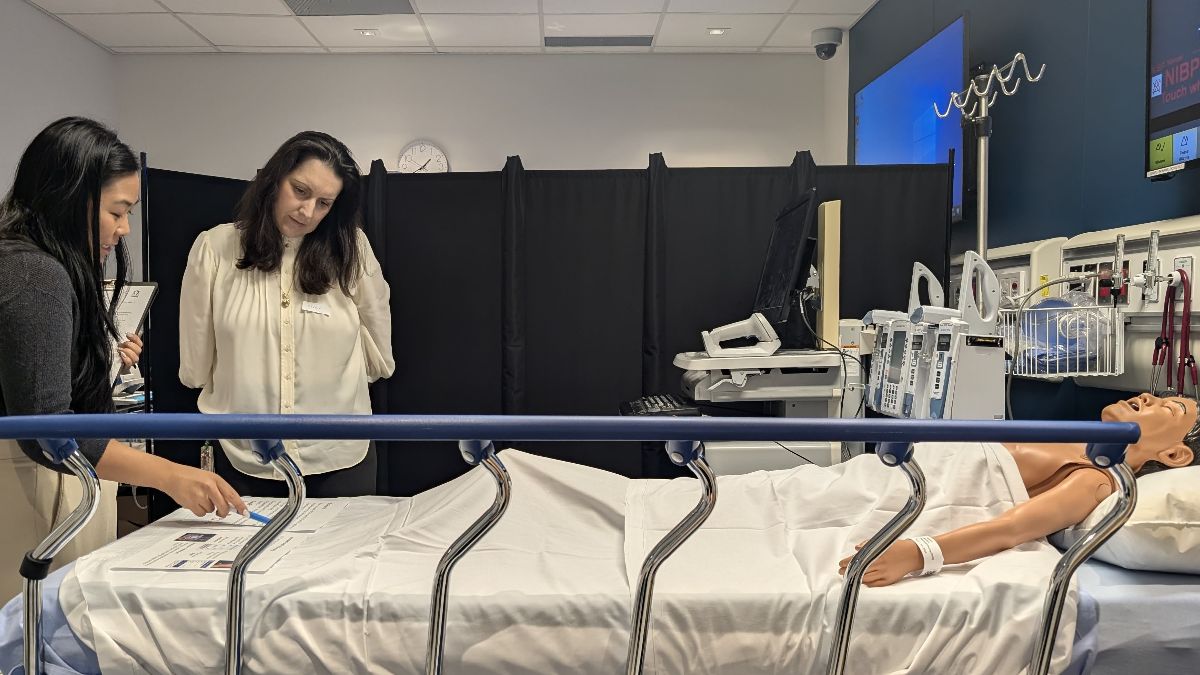Two people in a medical simulation room review a document beside a hospital bed with a mannequin.