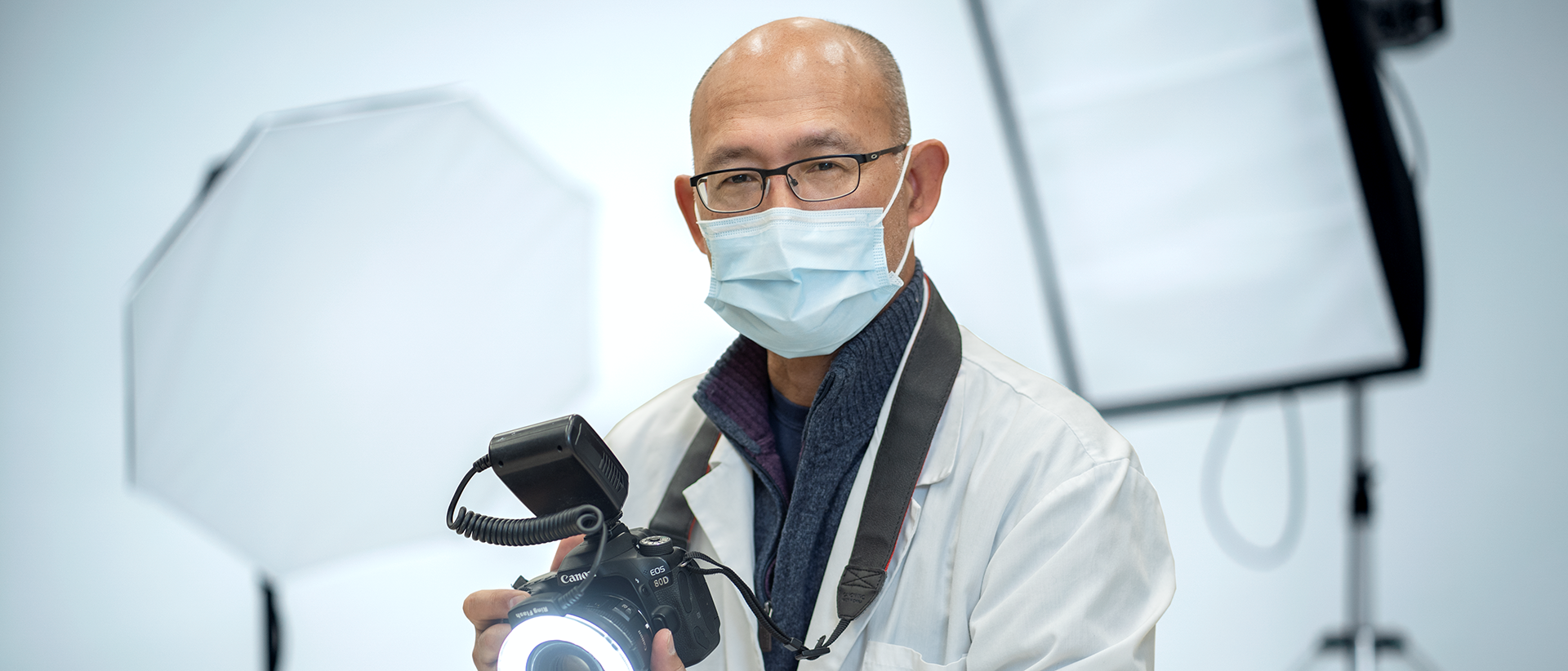 Dodge Baena wearing a mask and white lab coat while holding a camera. He is standing in front of a photoshoot set-up.