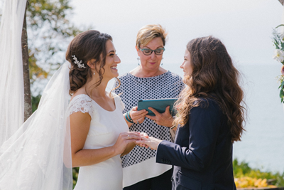 Taylor and Nicole Johnston wearing a wedding dress and suit hold hands at their outdoor wedding ceremony with officiant Sherry Murphy standing in the middle of them. 