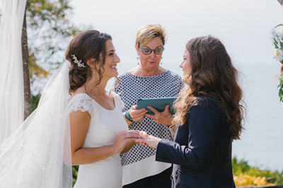 Taylor and Nicole Johnston wearing a wedding dress and suit hold hands at their outdoor wedding ceremony with officiant Sherry Murphy standing in the middle of them.