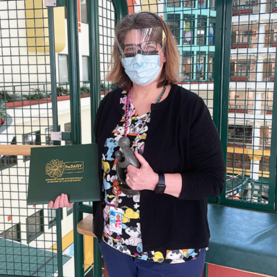 A woman wearing scrubs, a mask and face shield holds a small black statue and a green folder that reads Daisy Award