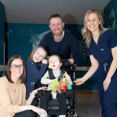 Three adults and a child surround another child seated in a wheelchair holding colorful toys in a hallway decorated with stars.