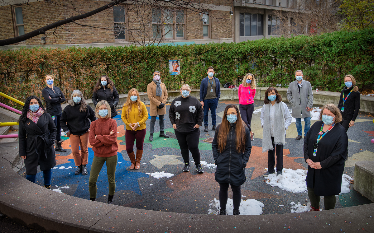 Group of staff stand together in a playground area with a hedge behind them. They are wearing masks and are physically distanced.