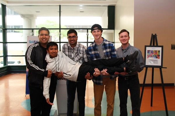 Four teen boys stand together smiling, holding up a fifth teen between them.