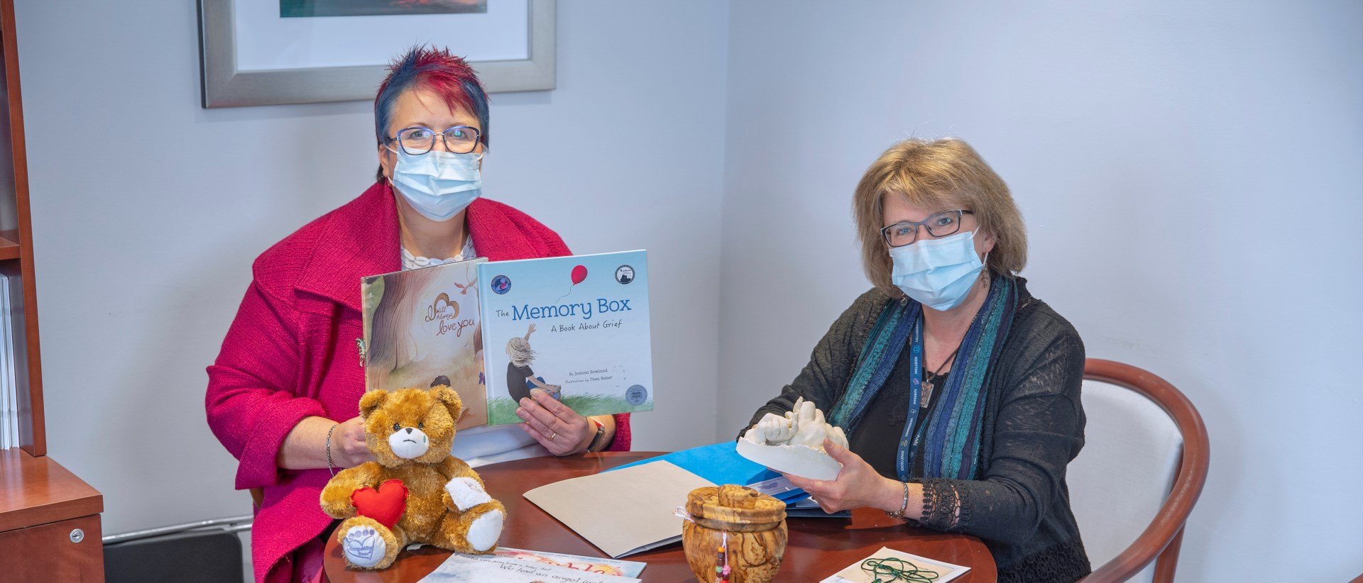 Lori Ives-Baine and Sheila Atkinson sitting at a small round table. Lori is holding a book that reads "The Memory Box, a Book About Grief". There are a number of items on the table, including a teddy bear, books, file folders, and a small urn with a colour beaded necklace hanging from it.