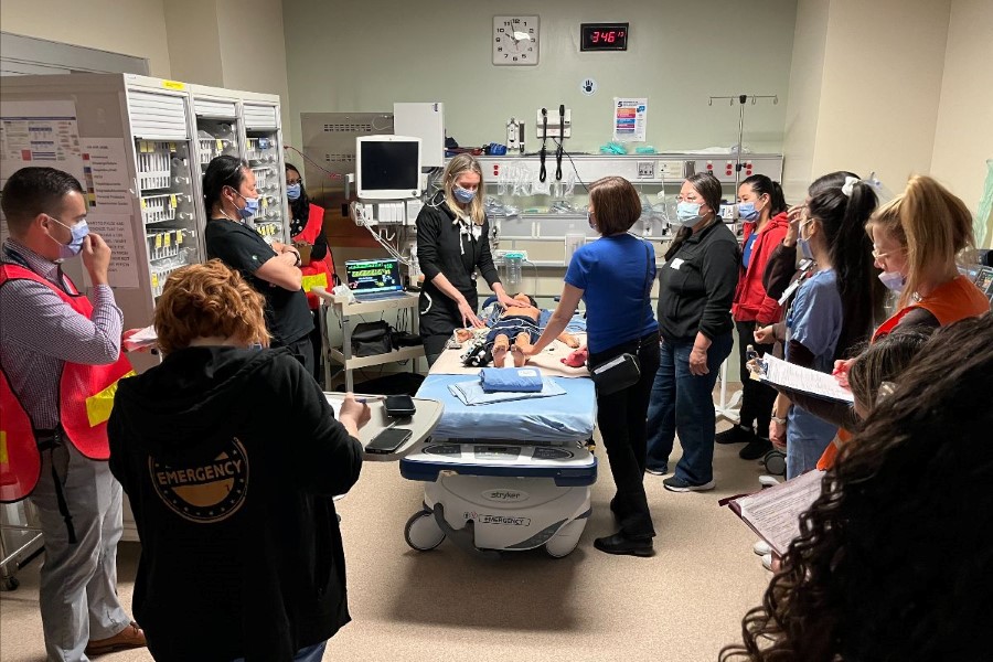 Medical staff gather around a hospital bed in an emergency room for a training session.