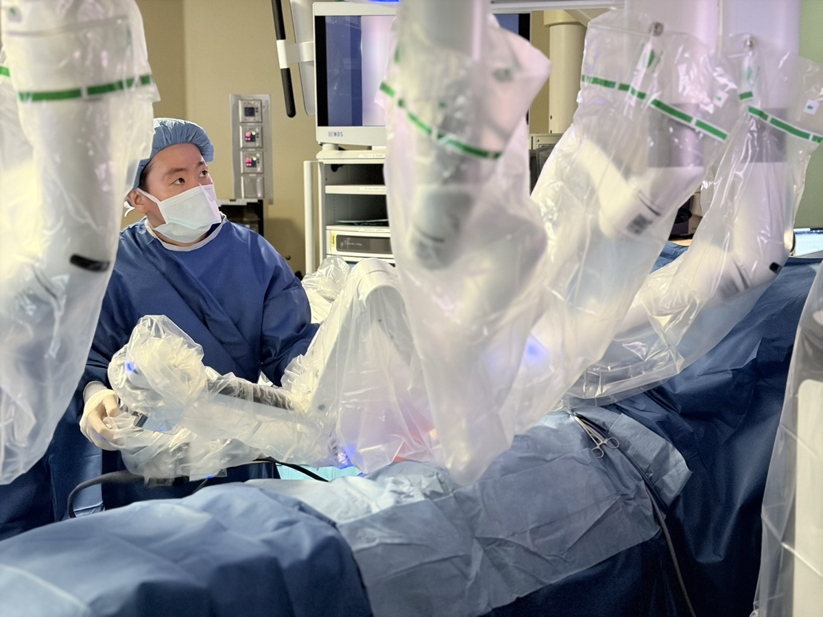 A man wearing blue scrubs, a cap and a mask positions the robot arms.