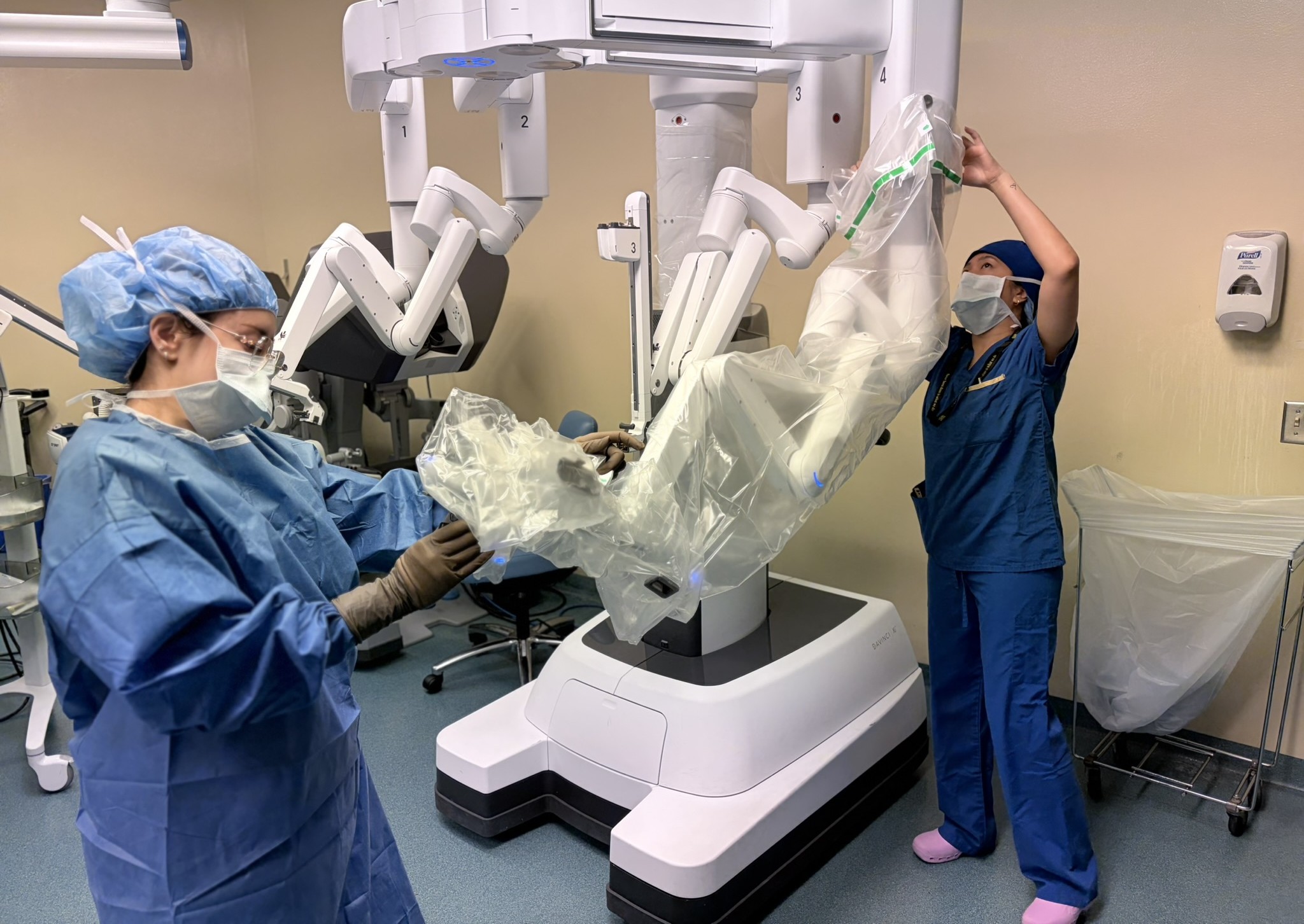 Two people in blue scrubs, masks and caps prepare different parts of a white robot.