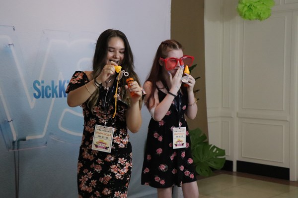 Two girls pose in front of a backdrop with silly accessories like big sunglasses and bubbles.