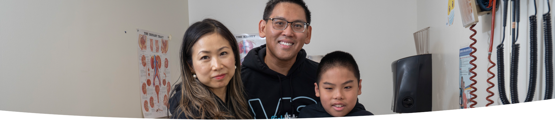 A family of three posing together for a photo at the Kidney Transplant Clinic. In the background, there are posters of kidney diagrams and medical equipment hanging from the walls.