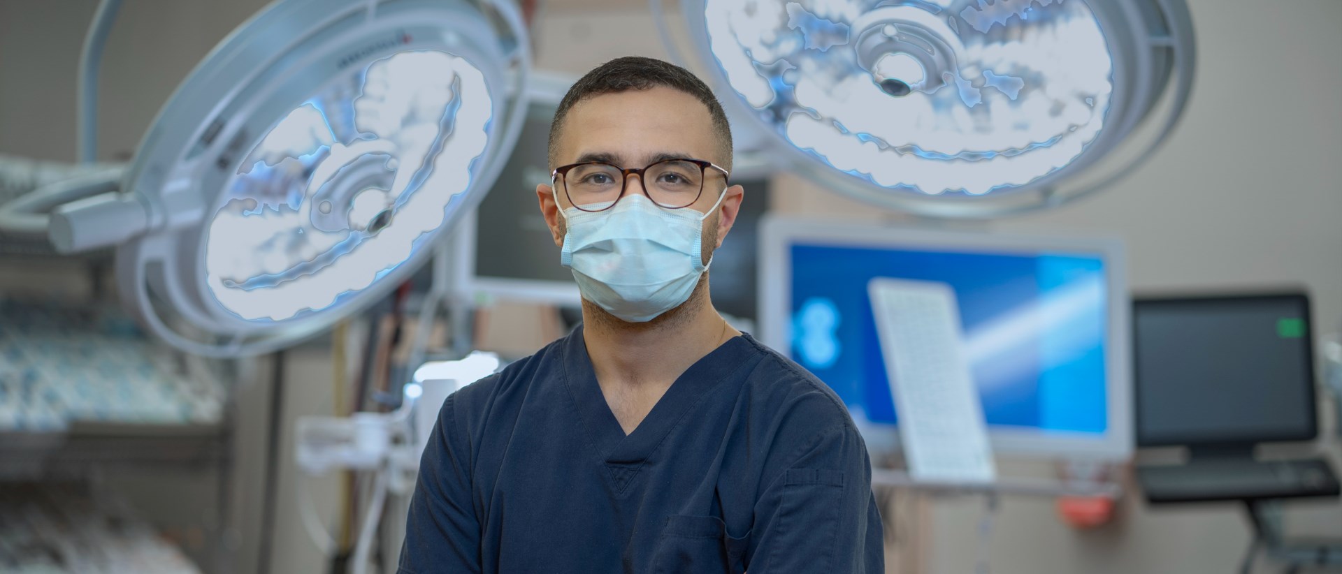 Dr. George Ibrahim in a mask standing in an operating room, with a variety of hospital equipment in the bacgkround.