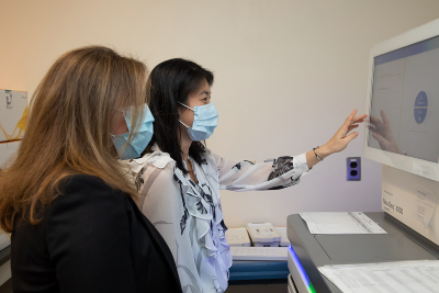 Dr. Kyriakopoulou and Dr. Yuki sitting in front of a machine that is labelled "NovaSeq 6000". Dr. Yuki is interacting with the screen on the machine.