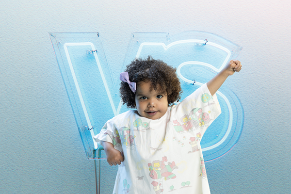 Toddler poses holding one arm pointing diagonally up. Behind them is a neon sign of the letters VS.