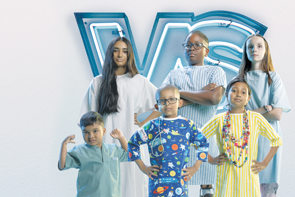 Six children wearing hospital gowns. The children are posing in front of a neon sign with the letters VS. Most of the children pose with their hands on hips or arms crossed. One child poses with arms raised.