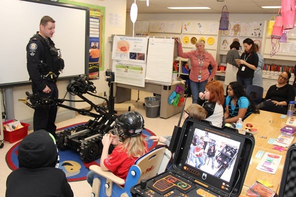 Police officer stands in a classroom with a robot, surrounded by children and teachers.