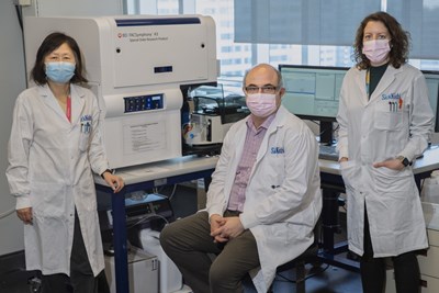 (left to right) Sheyun Zhao, John Moniakis, and Emily Reddy in masks and lab coats. There is a computer and lab equipment behind them.