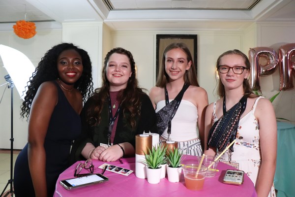 Four teenage girls stand side by side behind a table with drinks and decor. They are dressed in formal attire.