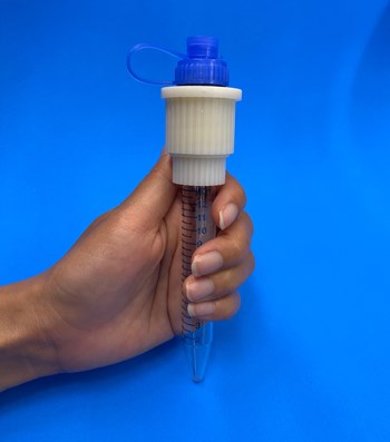 A hand holding a Capsule Shredder, a large tube that has measurement increments along the side and has a cylindrical adaptor and pop-top cap screwed to the top of the tube.