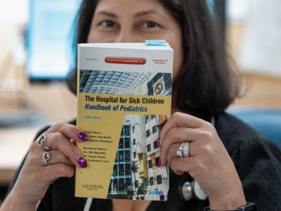 Dipchand holds a handbook in front of her face. The cover reads, "The Hospital for Sick Children Handbook of Pediatrics" with a yellow background and image of the hospital's Atrium.