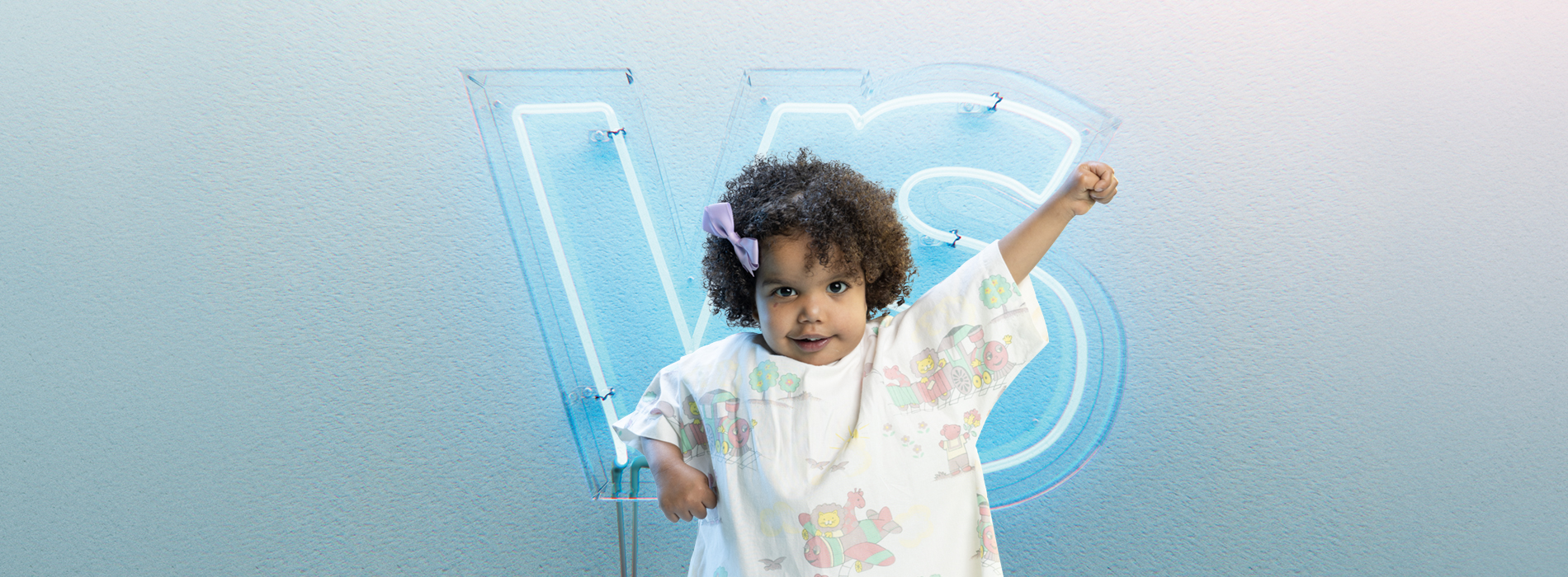 Young child wearing a hospital gown. She has one arm raised, standing in front of a neon sign with the letters VS.