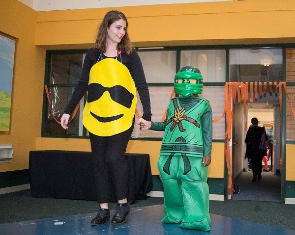 Boy dressed in a green ninja costume walks on stage. A woman wearing an emoji costume holds his hand.