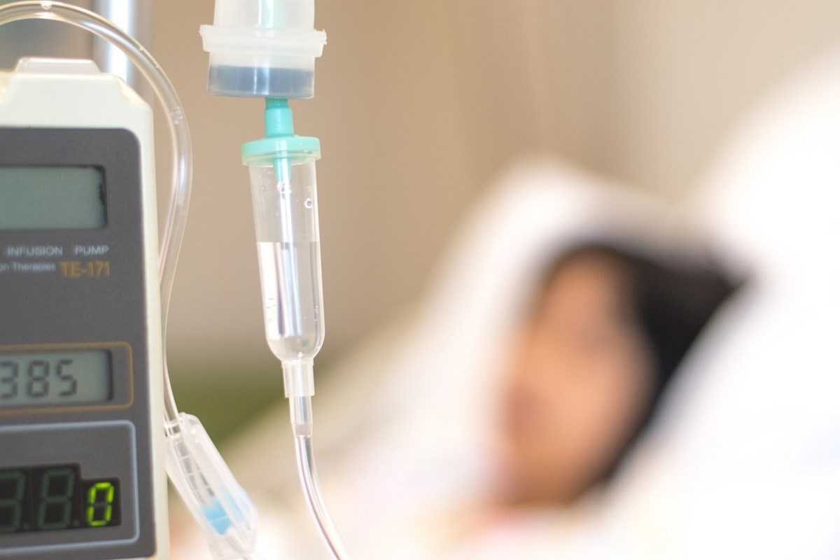 A closeup of an IV drip with a patient lying on a bed in the background  out-of-focus.