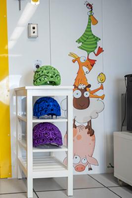 Three colourful OPM MEG helmets sitting on side table with shelves. There are cartoon animals painted on the wall next to the table.