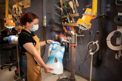 A person wearing a surgical mask and a brown workshop apron stands in a manufacturing lab holding a blue and white orthosis.