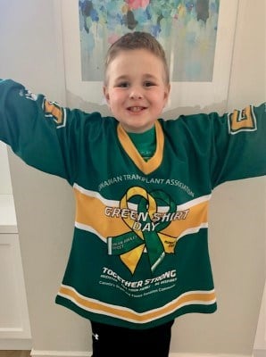 A young boy smiling with his arms raised in the air. He is wearing a green shirt with text that reads, "Canadian Transplant Association. Green Shirt Day. Together strong,"